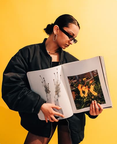 Person wearing sunglasses and a black jacket holding a magazine against a yellow background